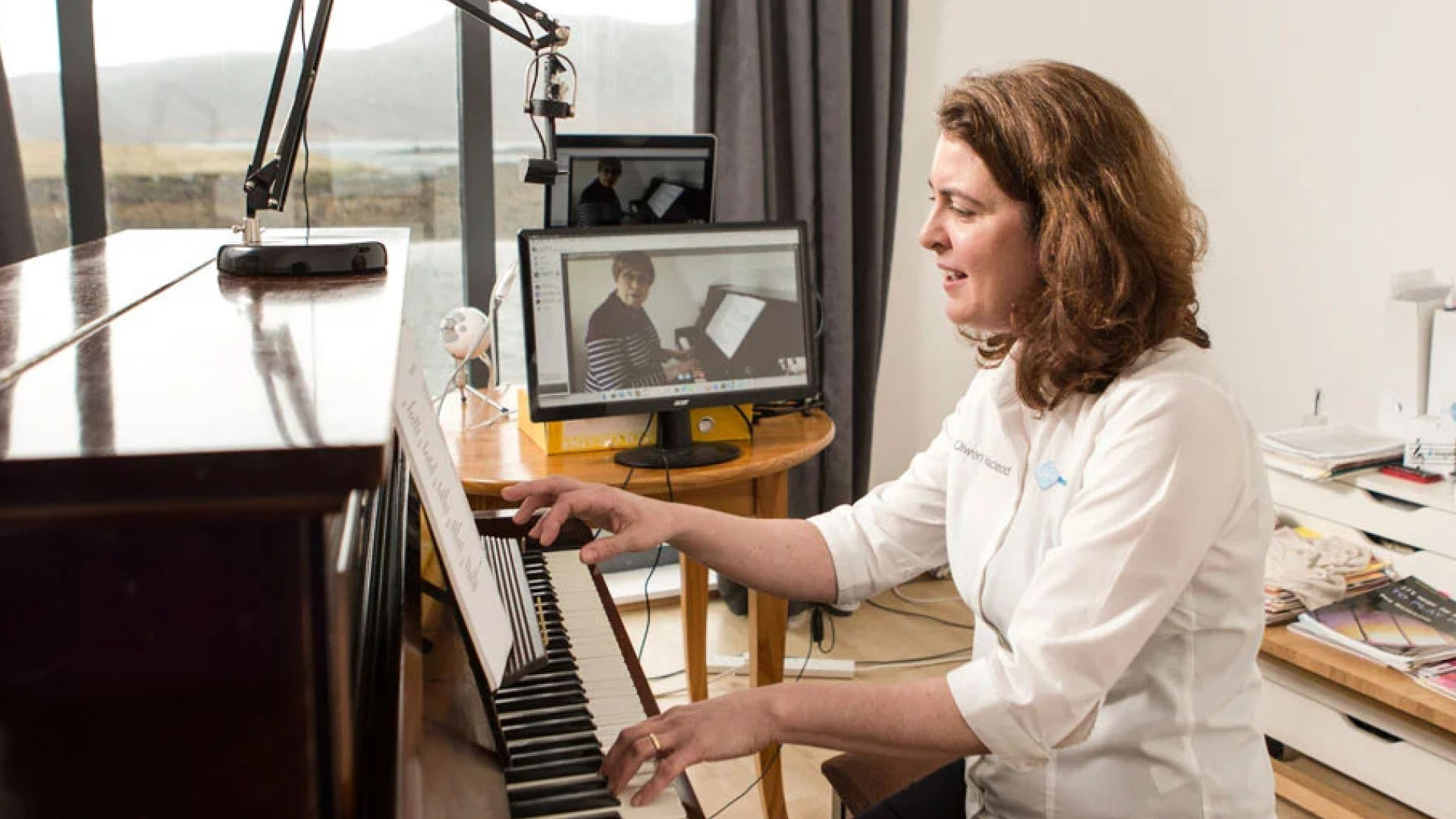 A teacher at the piano with a live lesson on her tablet